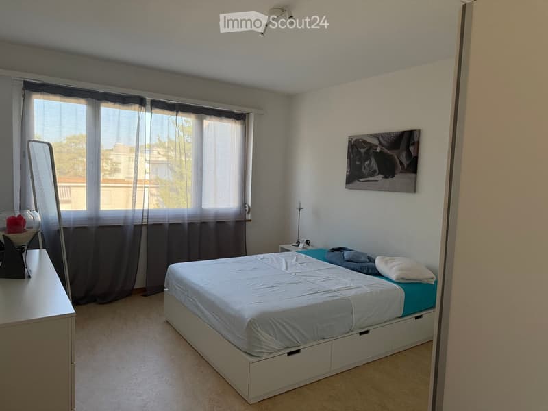 Modern bedroom with a white bed, nightstand, mirror, and picture frame on the wall. The room features large windows with dark curtains and a view of the outside.