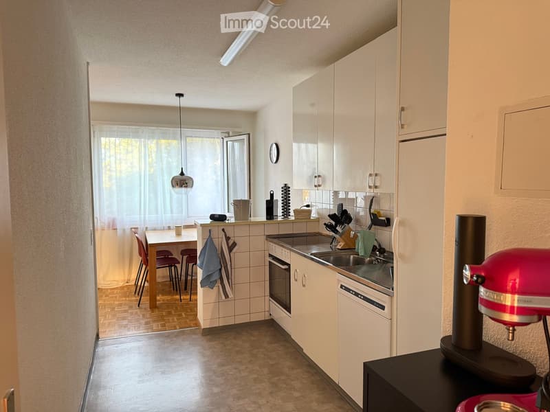 Modern kitchen with white cabinets, stainless steel appliances, a sink, oven, dishwasher, and a small dining area with a table and chairs. A red stand mixer is visible on the counter.