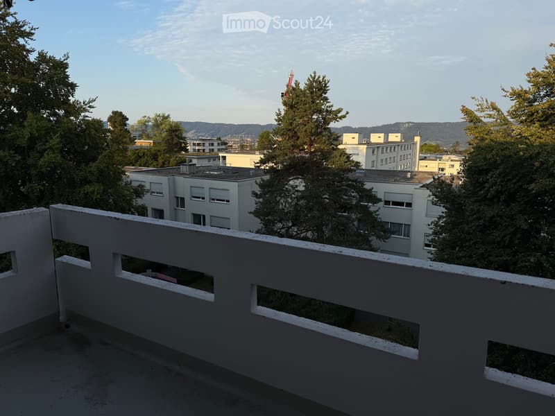 Balcony with white railing overlooking a cityscape with buildings, trees, and distant hills.