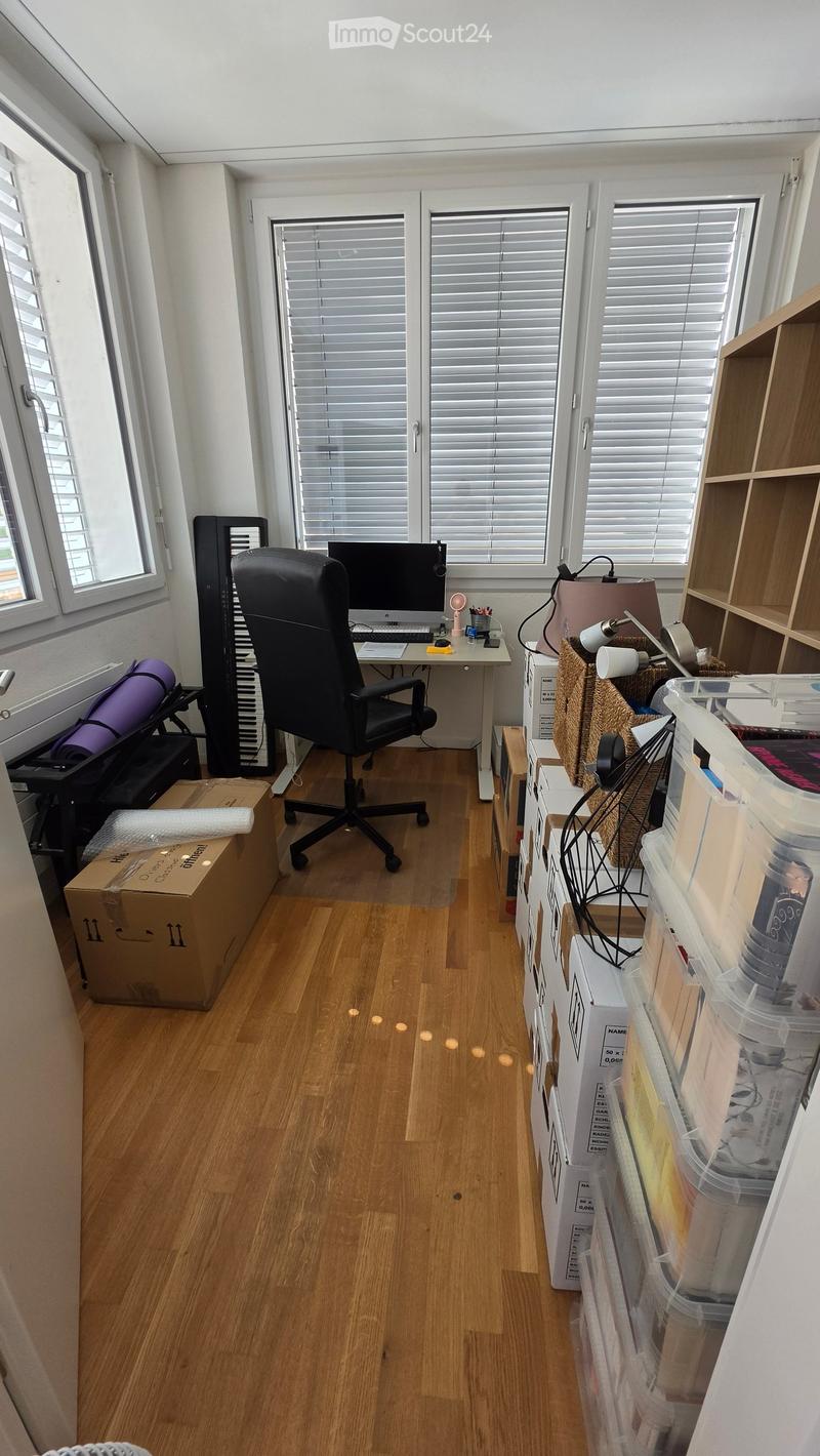 A well-lit office space with wooden flooring, large windows with blinds, and a desk setup including a computer, chair, and various storage items.