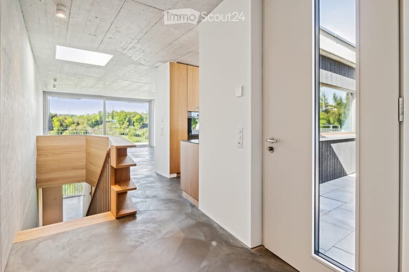 Modern, minimalist hallway with concrete floors, white walls, and large windows offering a view of greenery. Features a wooden staircase with open railings and built-in shelving, leading to an open-plan area with kitchen cabinets and a balcony.