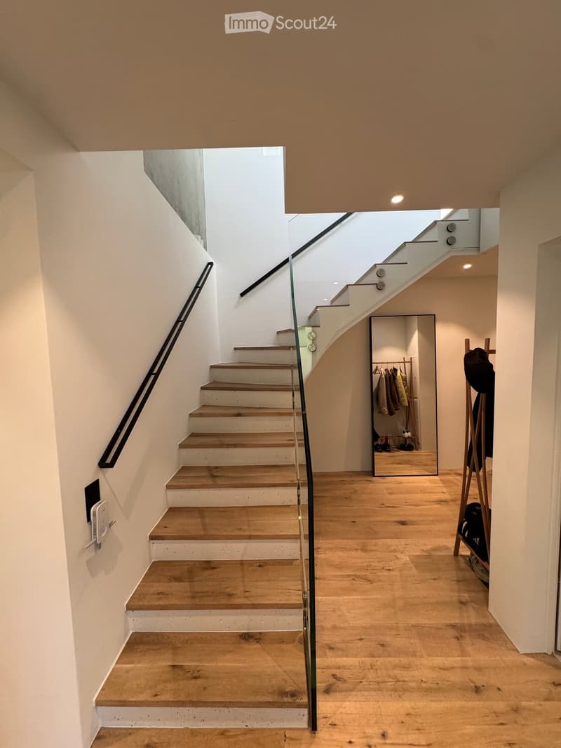 Modern hallway with wooden flooring, staircase with glass railing, coat rack, and mirror reflecting coats and shoes.