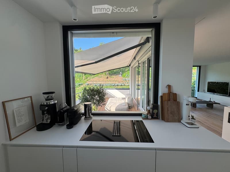 Modern kitchen with white countertops, stainless steel sink, coffee maker, and large window with retractable awning leading to outdoor seating area.