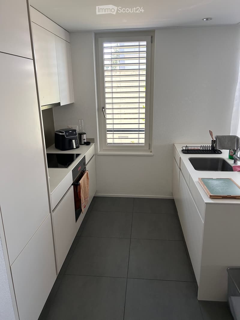 Modern kitchen with white cabinets, dark grey tiles, and stainless steel appliances. Features a coffee maker, oven, sink, dish rack, and cutting board.
