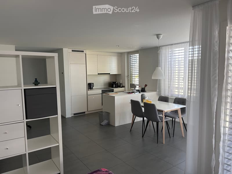 Modern kitchen with white cabinets, a central island, and a dining area with a table and chairs. The space features a sink, coffee maker, and a hanging lamp.