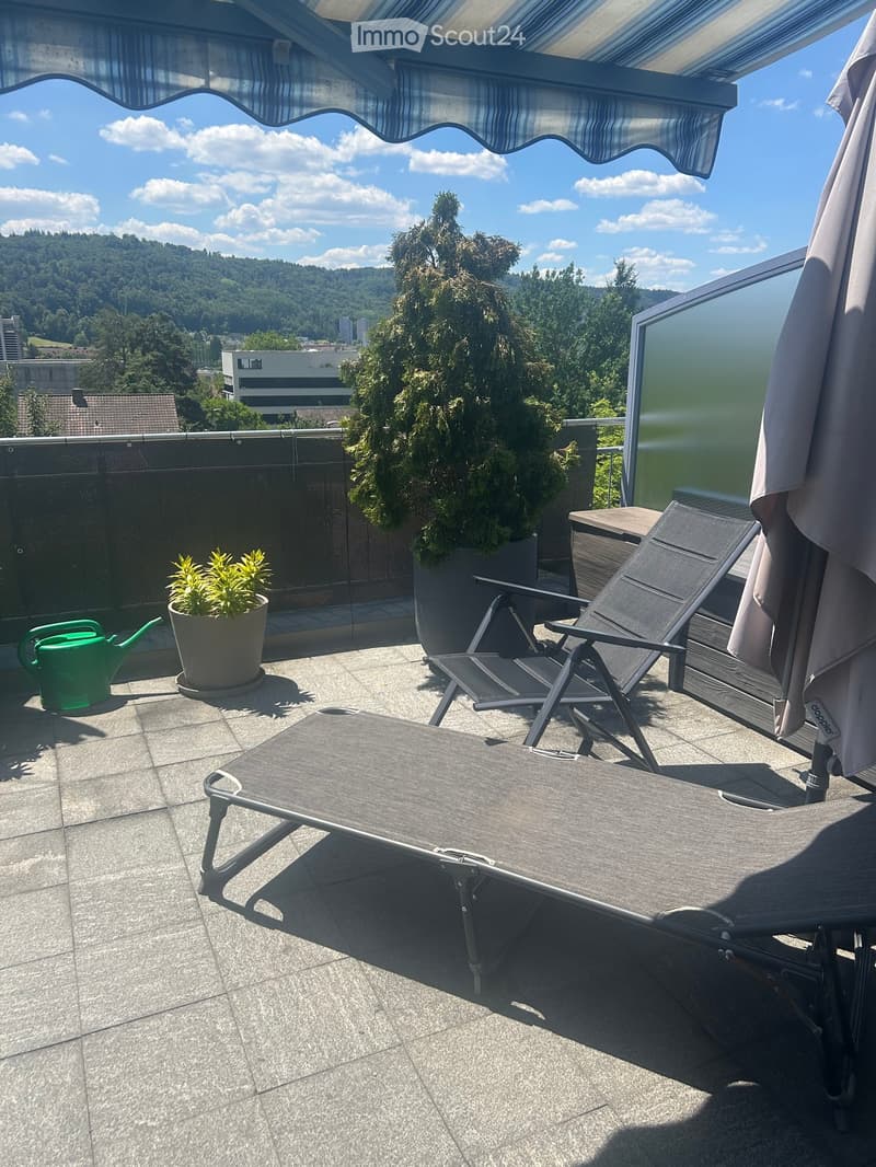 Balcony with lounge chairs, small table, umbrella, potted plants, and a scenic view of the city and hills.