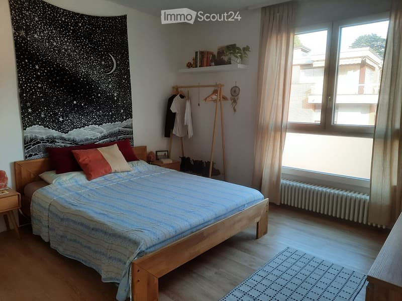 Cozy bedroom with a wooden bed, colorful pillows, a nightstand, a radiator, and a large window with curtains. A wooden hanger with clothes, a shelf with books and a plant, and a dreamcatcher are also visible.