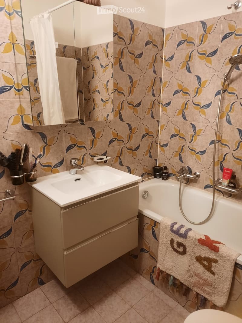 Modern bathroom with a white sink, beige cabinet, bathtub, and floral-patterned tiles. Includes a towel on the side of the tub and a shower curtain with a leaf design.