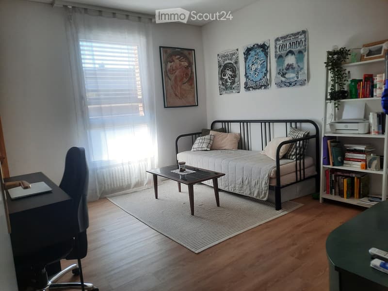 Cozy bedroom with a single bed, nightstand, desk, chair, and bookshelf. Features wooden flooring, large window with blinds, and decorative posters on the wall.