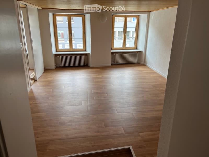 Empty room with wooden flooring, large windows, radiators, and a visible doorway leading to another space.