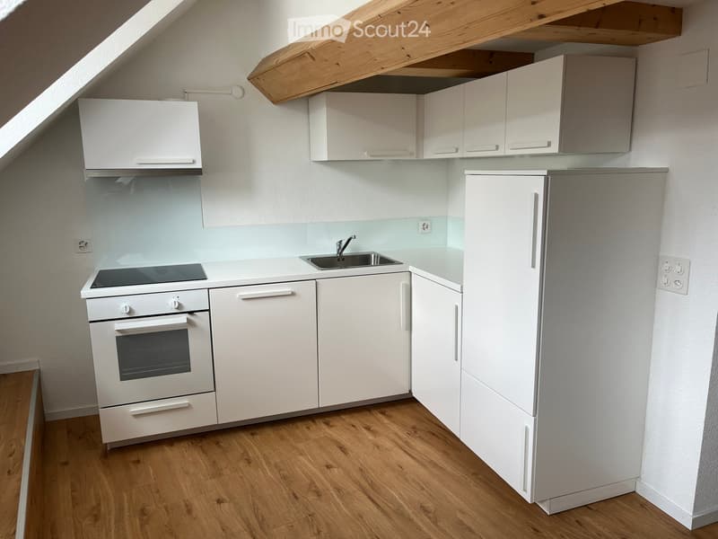 Modern kitchen with white cabinets, a refrigerator, an oven, and a sink, featuring wooden flooring and a slanted ceiling.