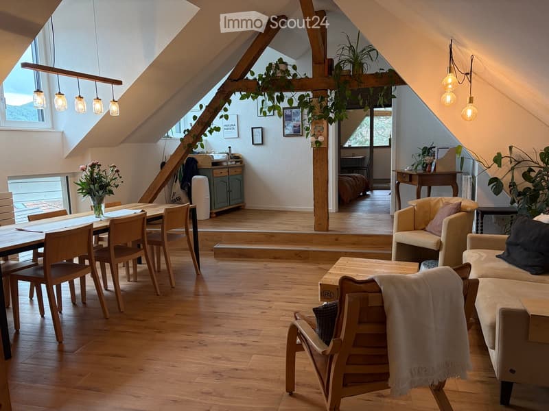 Bright, open living space with wooden floors, exposed wooden beams, and large windows. Features a sofa, coffee table, dining table with chairs, and decorative plants.
