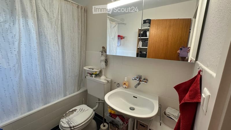 A compact bathroom with a toilet, sink, mirror, bathtub, and a wooden cabinet. A red towel hangs on the wall, and a shower curtain is visible in the background.