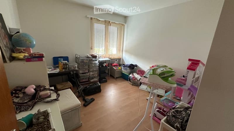 A cluttered room with wooden flooring, a desk with various items, a storage unit, an ironing board, a stool, a globe, and a birdcage.