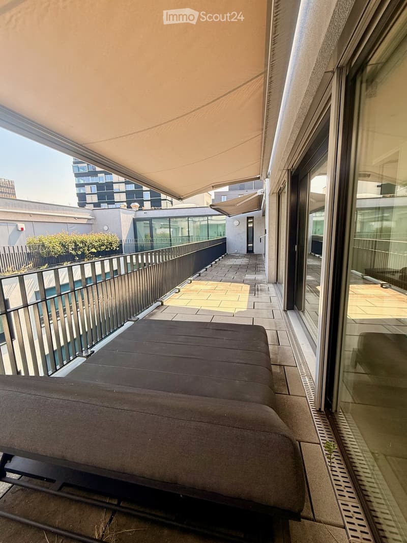 Balcony with a grey sofa, modern railing, and a retractable awning, overlooking a cityscape with modern buildings.