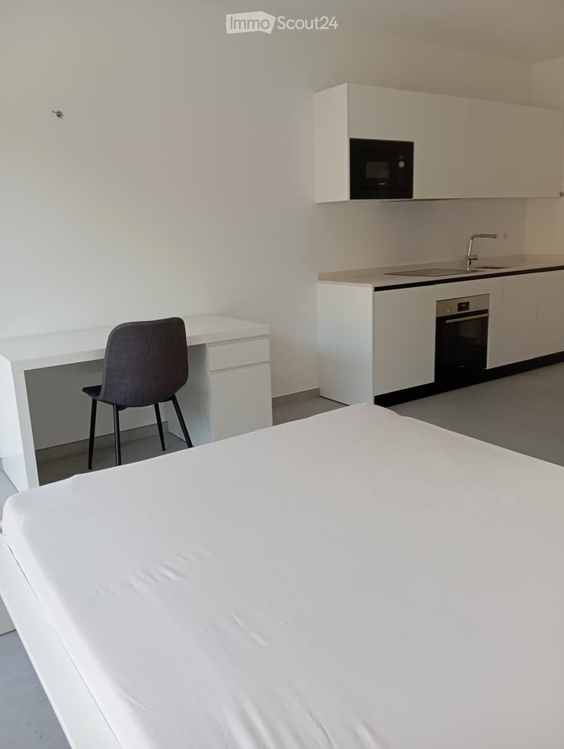 Modern studio apartment with a bed, desk, chair, and a compact kitchen area featuring a microwave, oven, and sink.