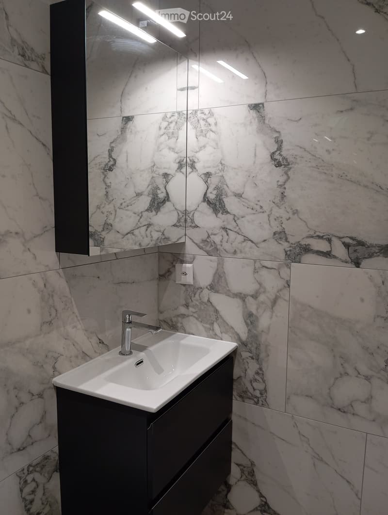 Modern bathroom with marble walls, a white sink, a chrome faucet, and a large mirror with built-in lighting.