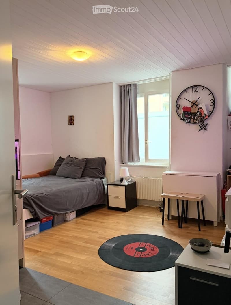 Cozy bedroom with wooden flooring, slanted ceiling, large window with curtains, bed with pillows and blanket, nightstand with lamp, small table, stool, vinyl record, and decorative clock.