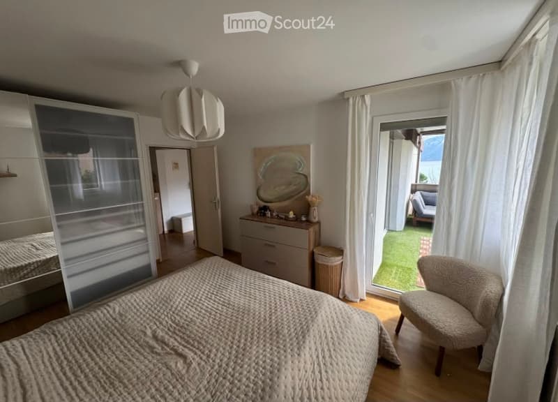 Modern bedroom with wooden flooring, large window with white curtains, bed with textured duvet, armchair, dresser with drawers, mirror, and a decorative art piece on the wall.