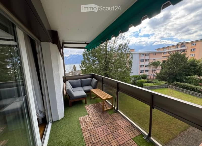 Balcony with a grey sofa, wooden coffee table, green artificial grass, and a brick-tiled section. Offers a scenic view of mountains and surrounding greenery.