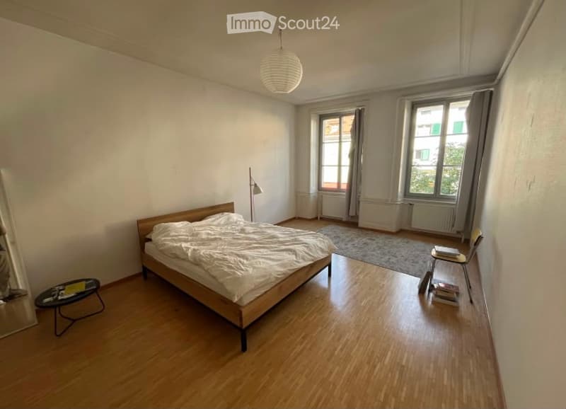 A modern, well-lit bedroom with wooden flooring, a large bed with white bedding, a small round table with books, a chair, and a floor lamp near the window.