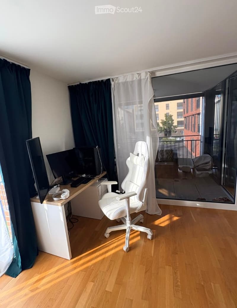 Modern, well-lit room with wooden flooring, a white ergonomic chair, a desk with a computer screen and keyboard, and large windows leading to a balcony.