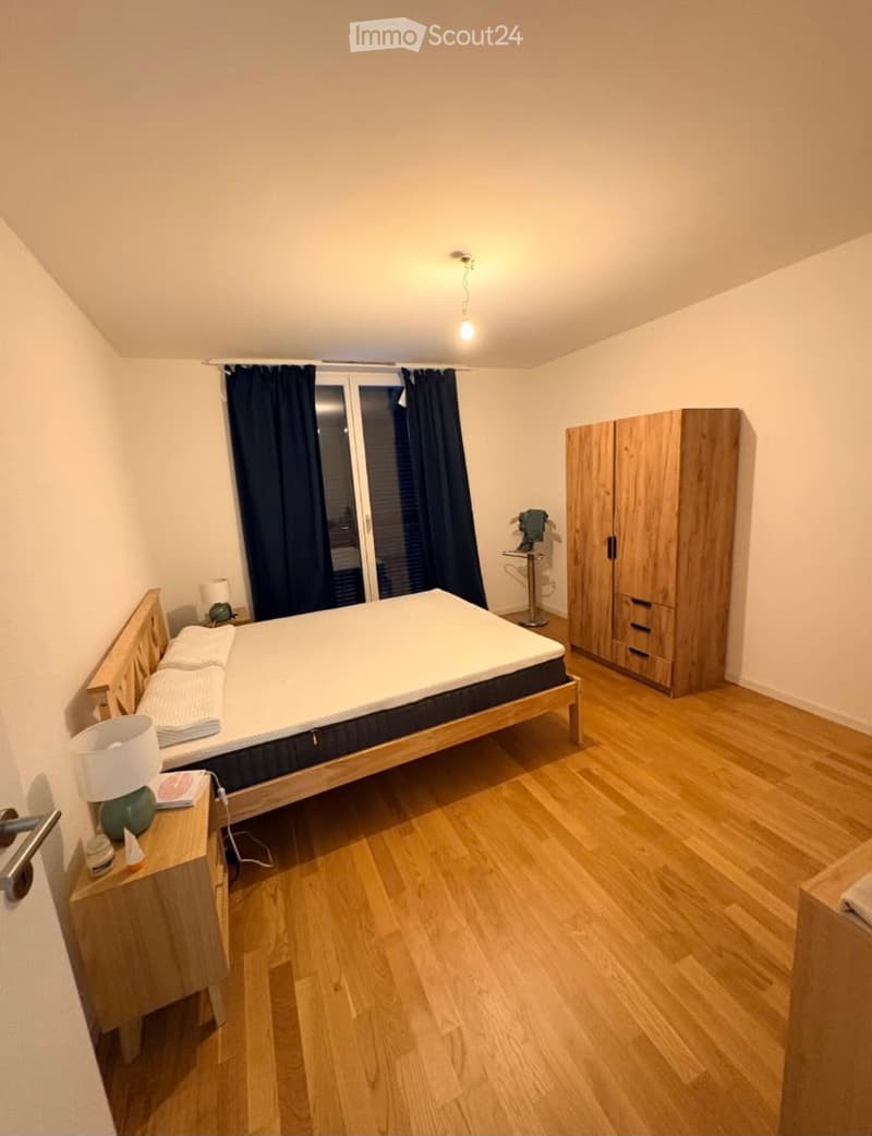 Cozy bedroom with wooden flooring, a double bed with white bedding, a wooden nightstand with a lamp, a wardrobe, and a large window with dark curtains.