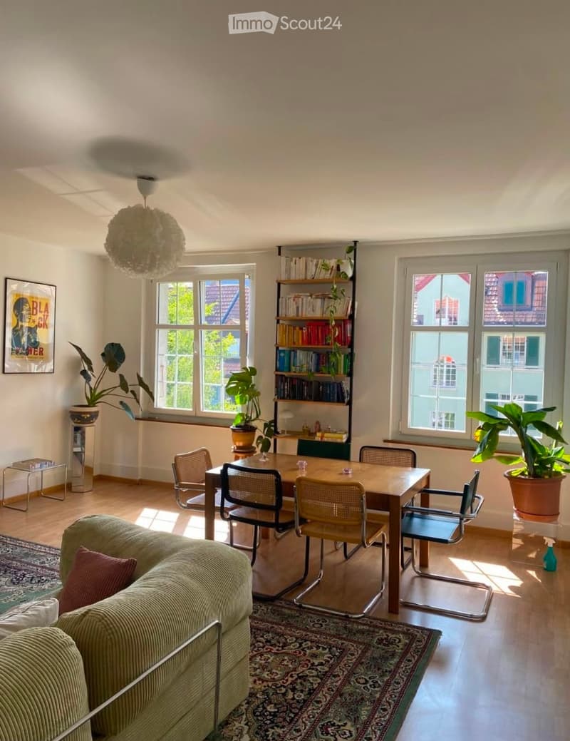 Bright living room with wooden flooring, large windows, a sofa, dining table with chairs, bookshelf, and potted plants.