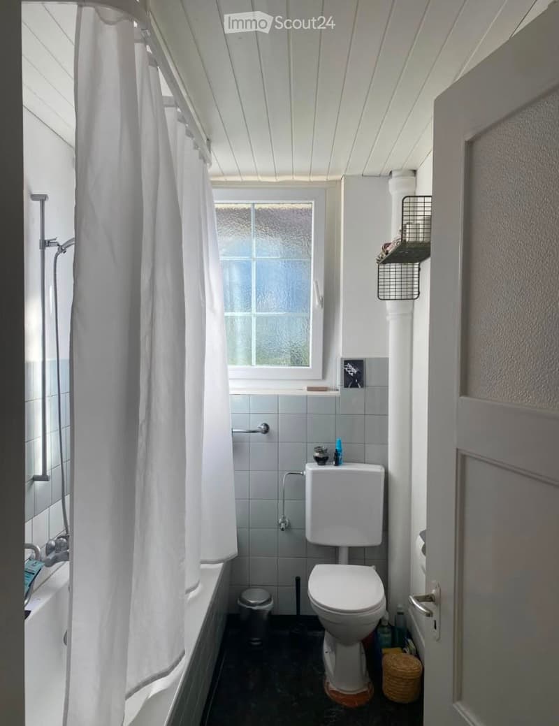 A small bathroom with a bathtub, toilet, shower curtain, and a window above the bathtub. The walls are tiled, and there is a shelf on the wall holding various items.