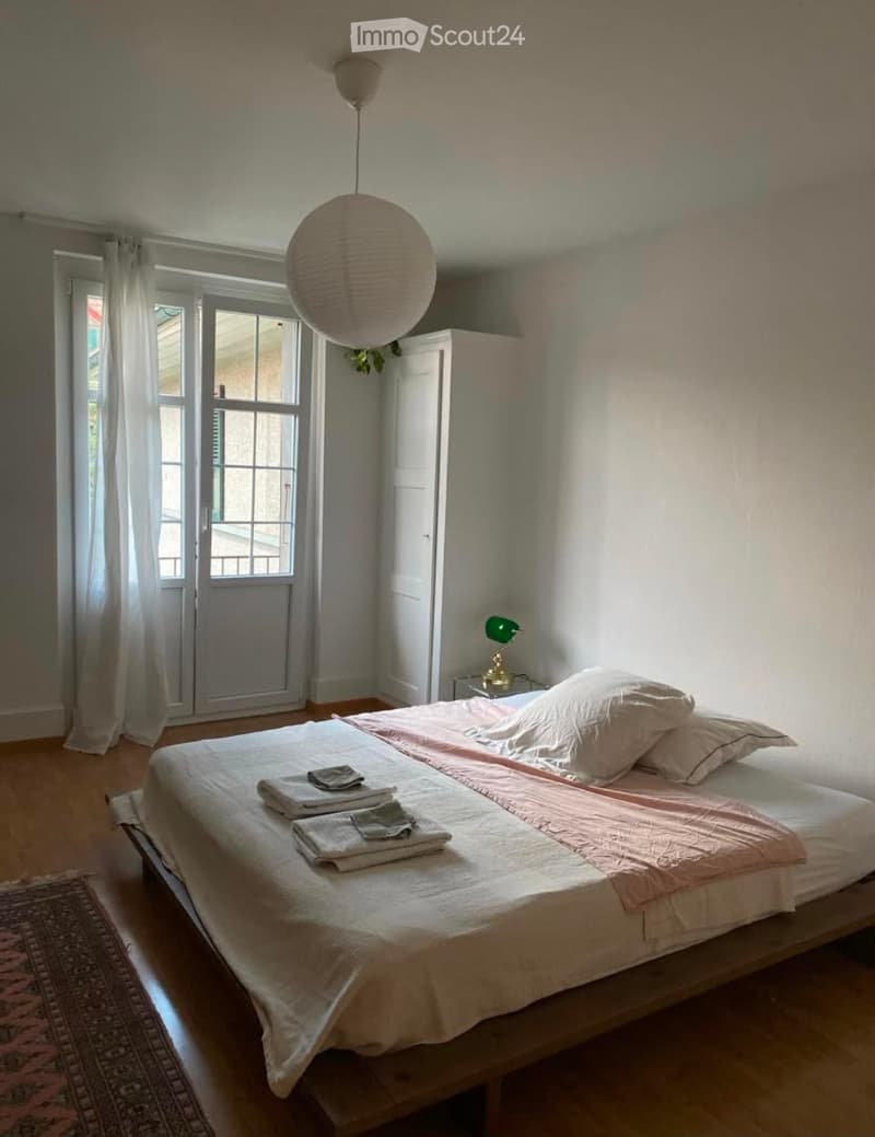 Cozy bedroom with a large bed, nightstand, and a hanging paper lamp. The room features wooden flooring, a window with white curtains, and a small rug near the bed.