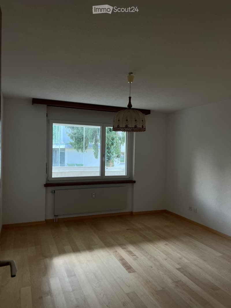 Empty room with wooden flooring, large window, radiator, and a hanging light fixture.