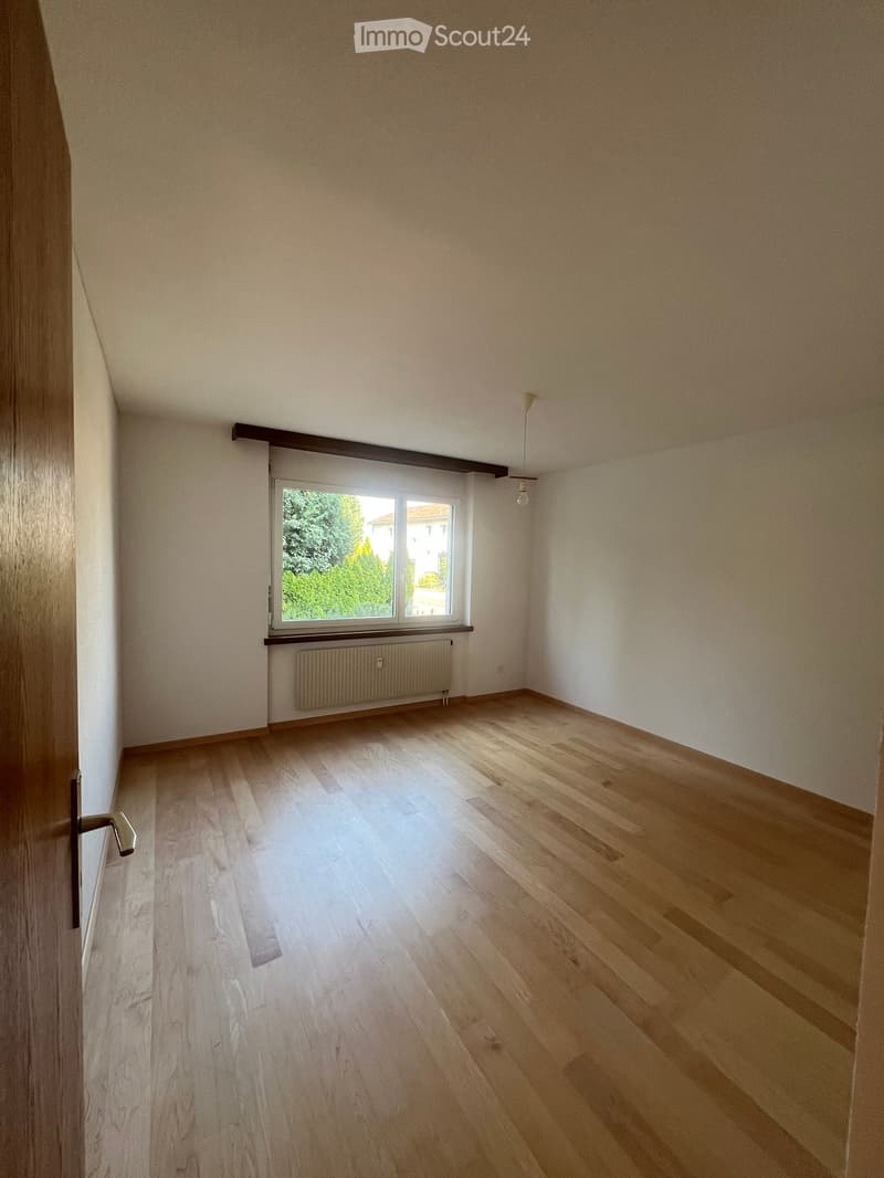 Empty room with wooden flooring, white walls, large window with view of greenery, radiator, and pendant light fixture.