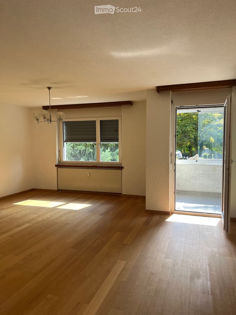 Empty room with wooden flooring, large window with blinds, radiator, and an open sliding door leading to a balcony or outdoor area.