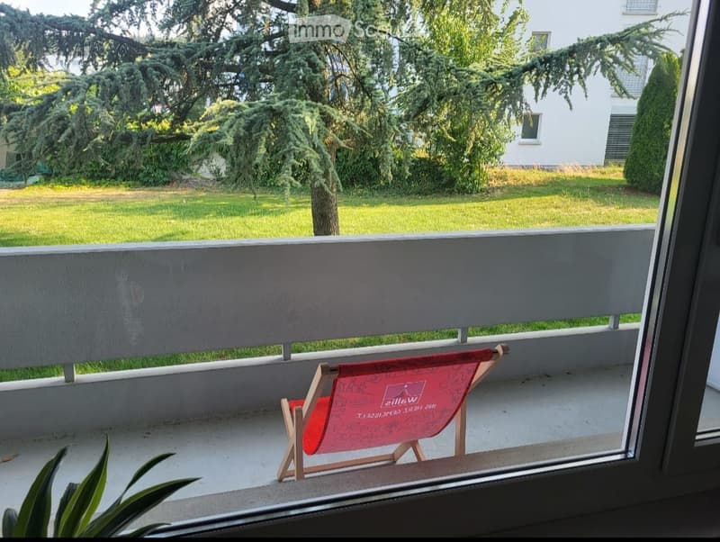 Balcony with a small red folding chair, overlooking a green lawn and trees.