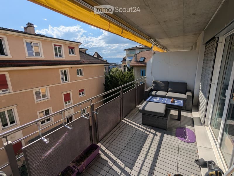 Balcony with wooden decking, metal railing, sofa, table, stool, rug, and bicycle.
