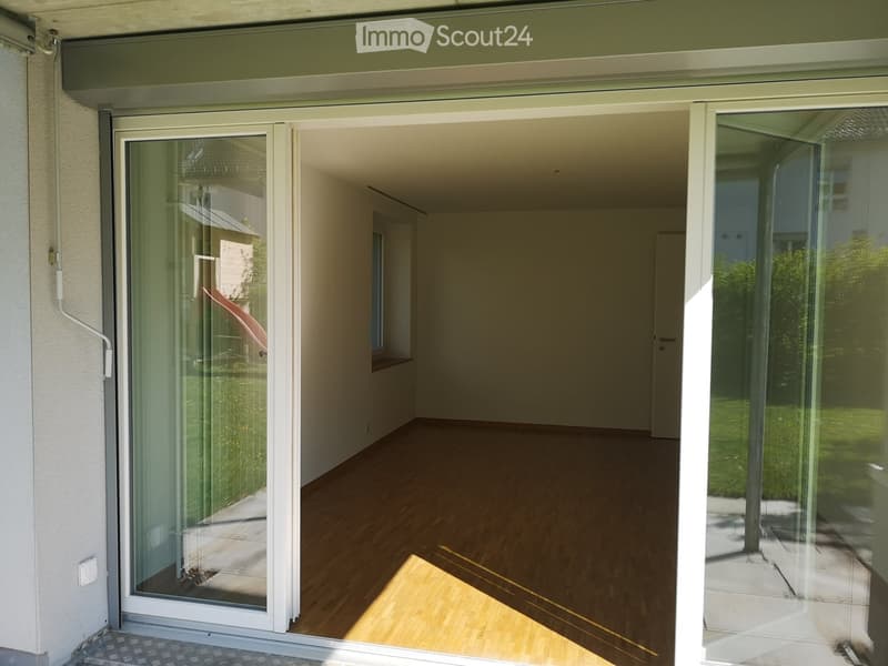 Empty room with wooden flooring, large glass sliding doors leading to an outdoor area, and visible sunlight streaming in.