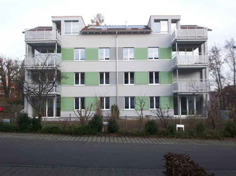 Modern multi-story apartment building with grey and green facade, multiple balconies, and surrounding greenery.