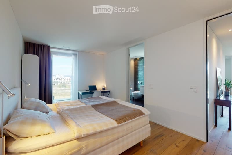 Modern bedroom with a large bed, wooden flooring, a desk with a laptop, a small table with a plant, and a mirror on the wall. The room has large windows with curtains and a view of the city.