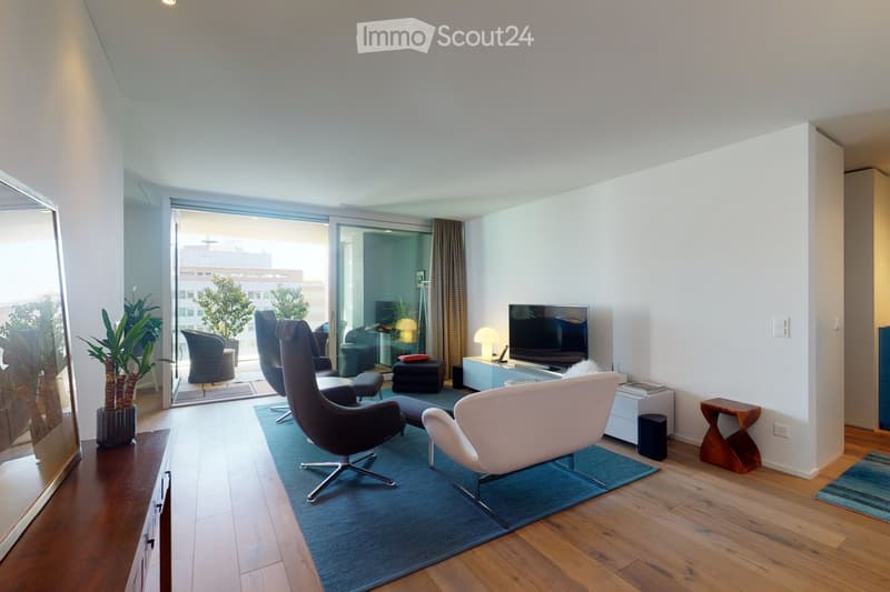 Modern living room with wooden flooring, large sliding glass doors leading to a balcony, a blue rug, a white armchair, a black leather chair, a small wooden side table, a television, and a lamp.