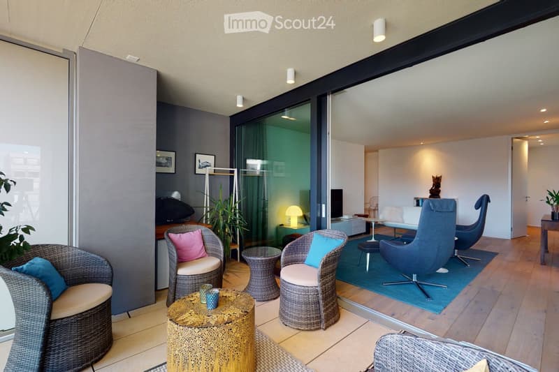A modern, well-lit living room with a mix of wicker and contemporary furniture, including a sofa, chairs, tables, and a lamp. The space features large windows with a view and is decorated with plants and artwork.