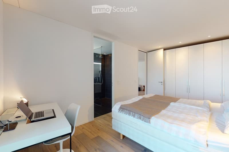 Modern bedroom with a white bed, laptop on a desk, white wardrobe, and a visible adjacent bathroom with dark tiles.