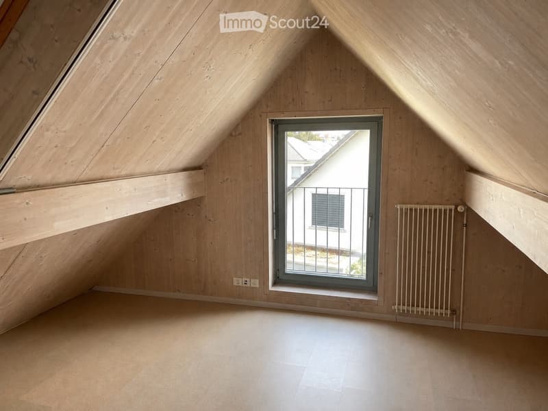 Empty attic room with wooden walls and ceiling, large window, radiator, and visible electrical outlets.