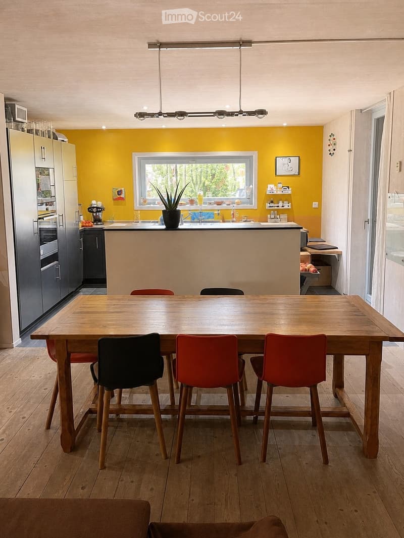 Bright kitchen with a yellow accent wall, wooden dining table, and modern appliances. Features include a bar counter, stools, and a potted plant on the counter.