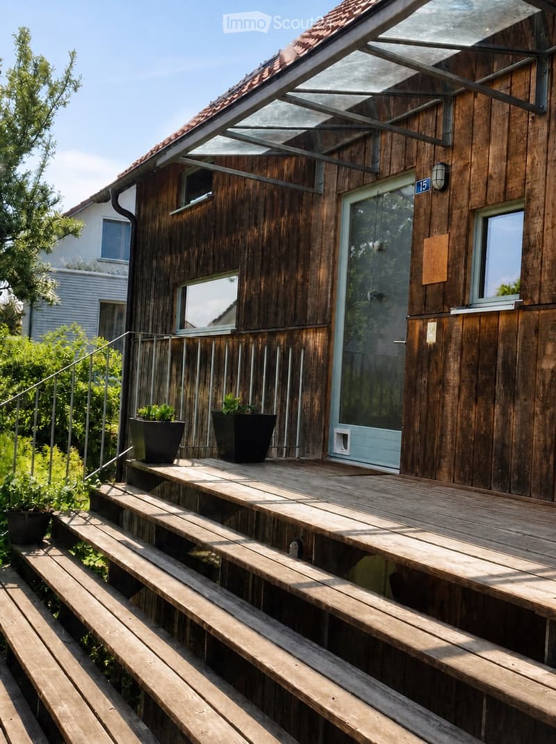 Wooden balcony with steps leading up, featuring potted plants and a modern glass door entrance to the building.