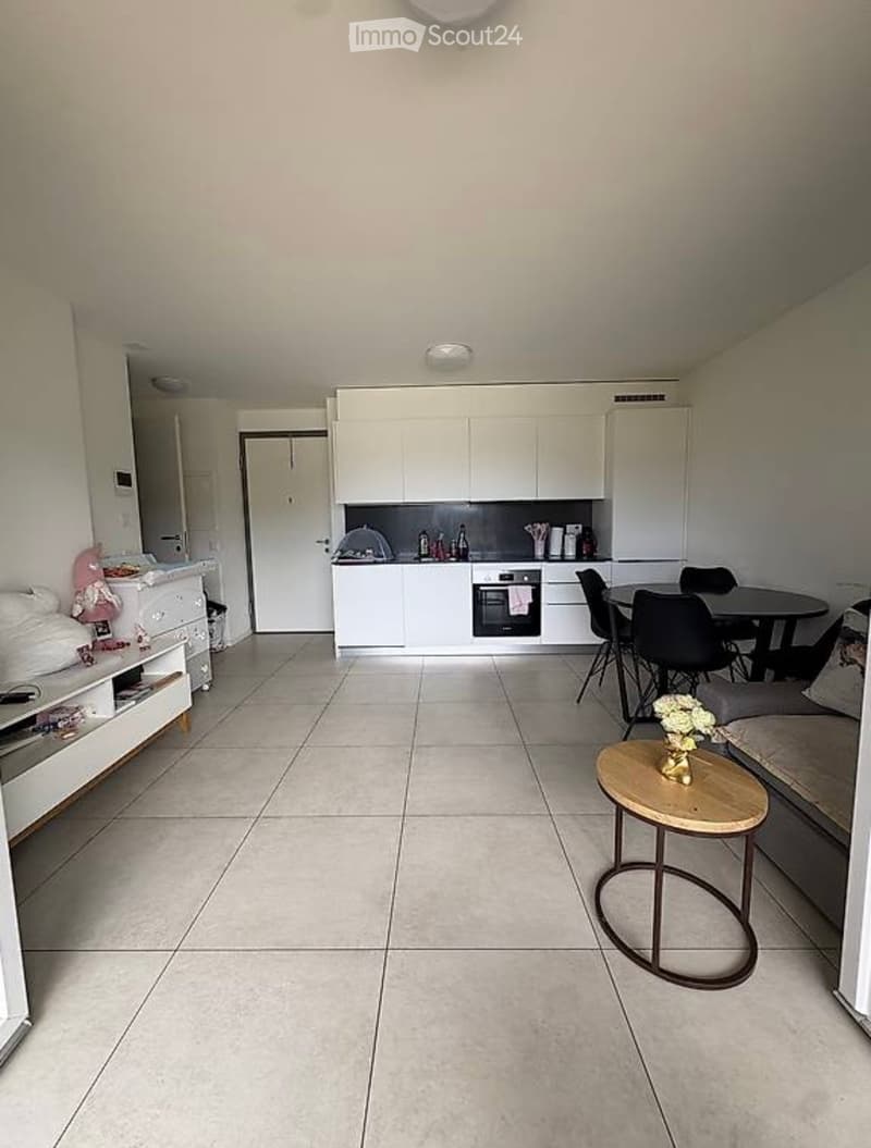 A modern, open-plan living and dining area with a kitchen. The space features a sofa, a small round table with a vase of flowers, a dining table with chairs, and a kitchen area with white cabinets and an oven.