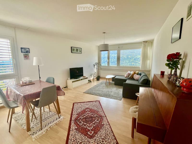 A modern living room with a sofa, coffee table, dining table, chairs, television, floor lamp, and decorative vases. The room features large windows with a scenic view, wooden flooring, and patterned rugs.