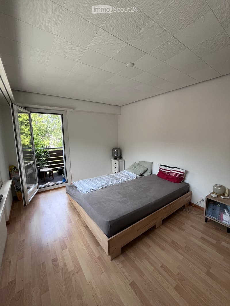 Cozy bedroom with wooden flooring, a single bed with grey bedding, nightstand, and an open sliding door leading to a balcony.