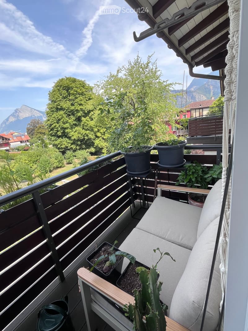 Balcony with a grey sofa, small wooden table, and several potted plants, overlooking a scenic mountain view.