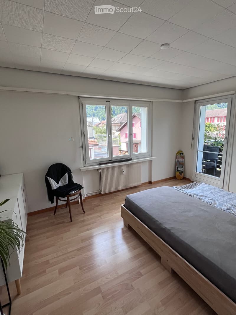 Bright bedroom with wooden flooring, large windows offering a view of the neighborhood, a bed with grey bedding, a wooden chair with a jacket draped over it, a skateboard leaning against the wall, and a small plant near the window.