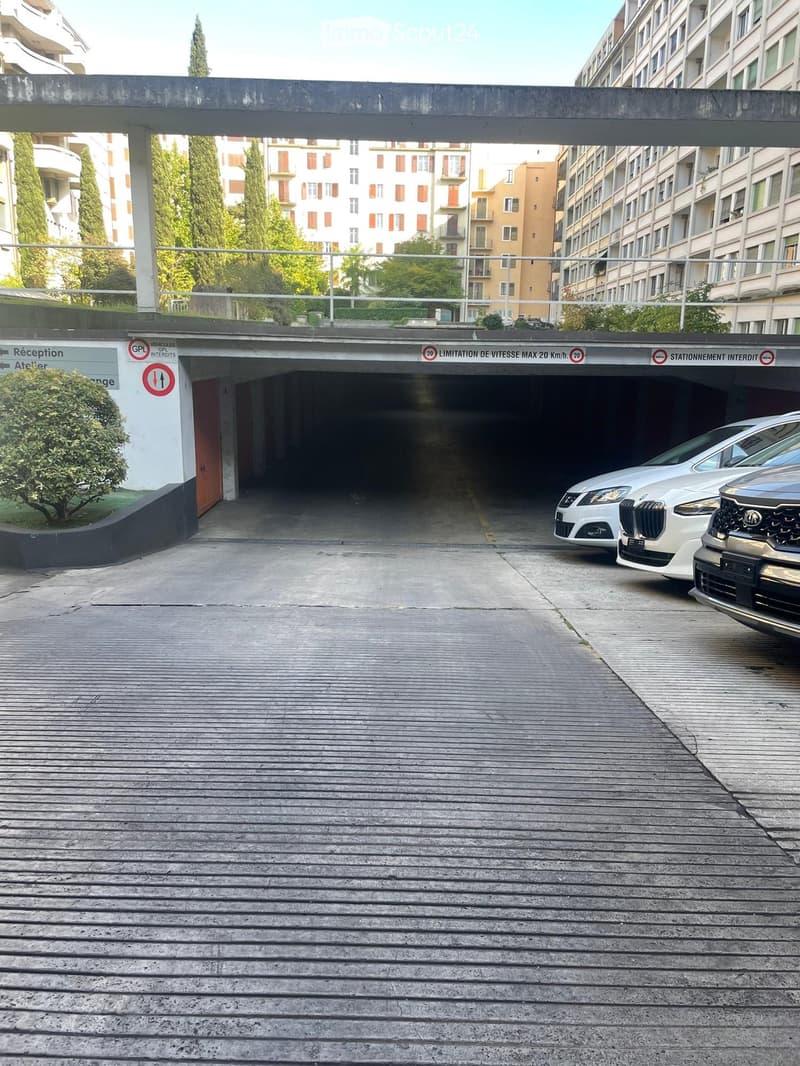 Entrance to a multi-story parking garage with a covered walkway leading to the entrance, several parked cars visible, and surrounding buildings in the background.